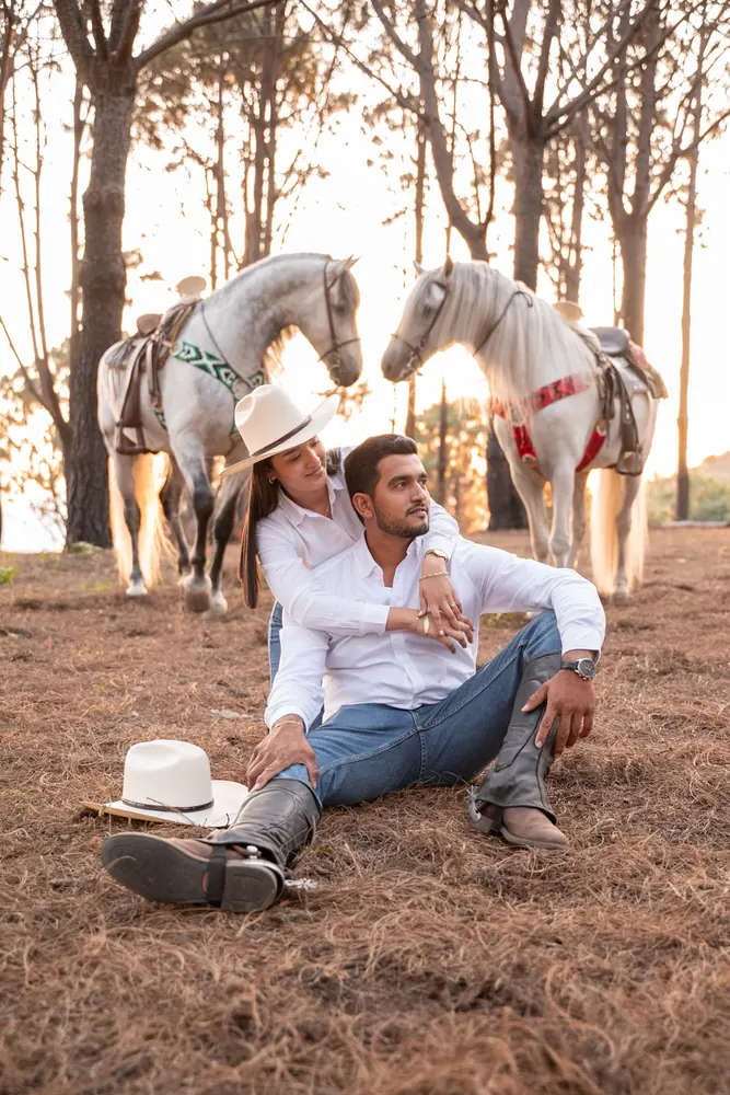 Jorge sentado en el pasto y Estephany abrazándolo, con dos caballos blancos detrás